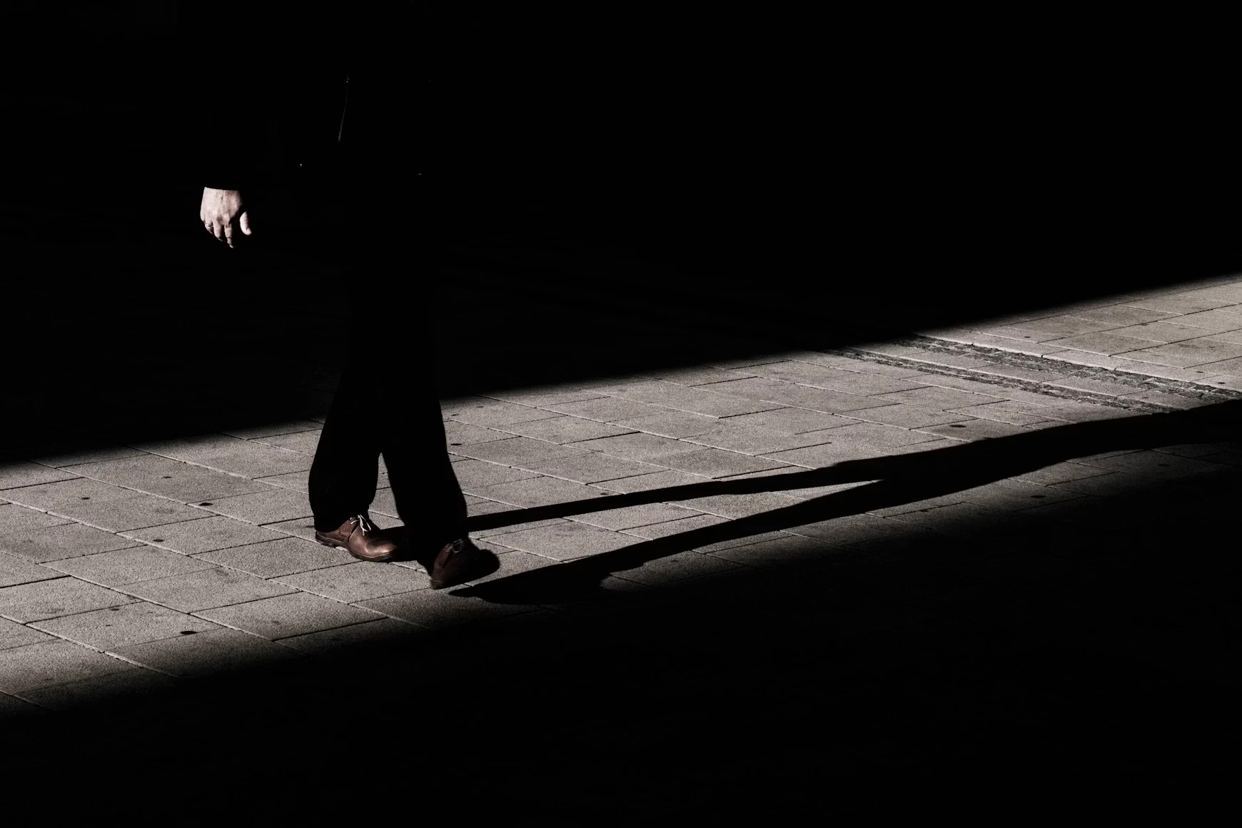 A lone figure in dark clothing walks through a shaft of light at night, a long shadow trailing across the empty pavement behind him.